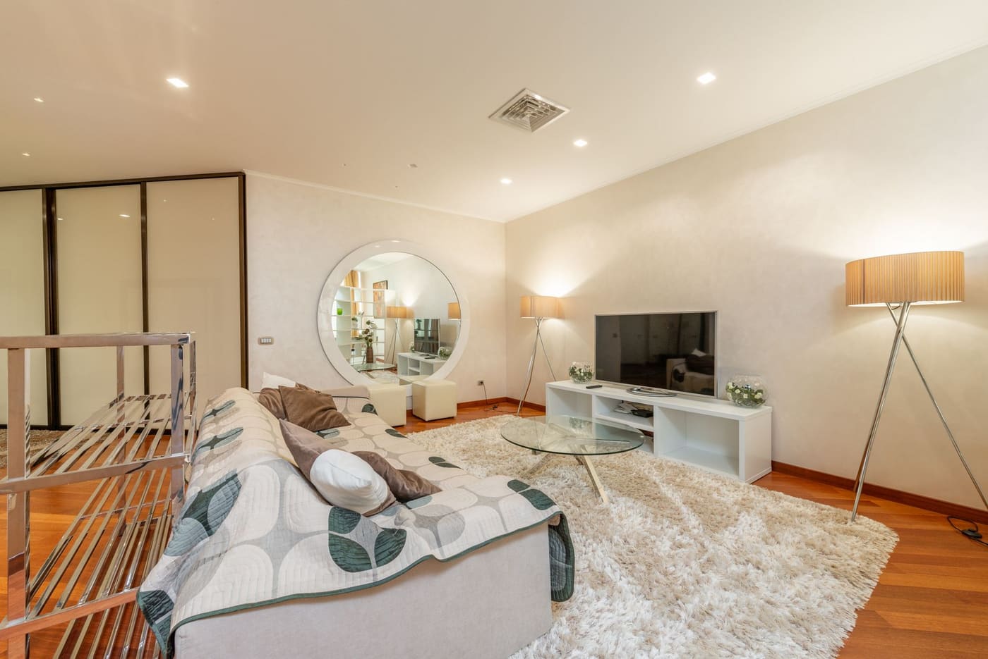 A bright apartment living room with a sofa, large rug, floor lamps and a wall-mounted TV