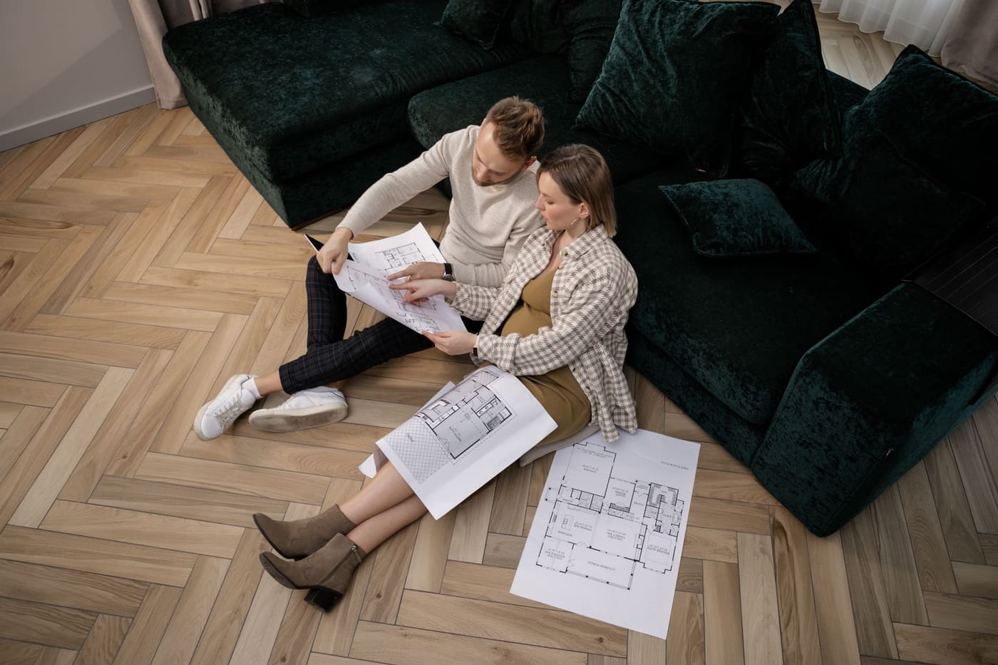 A couple sitting on a wooden floor beside a sofa, looking over printed floor plans
