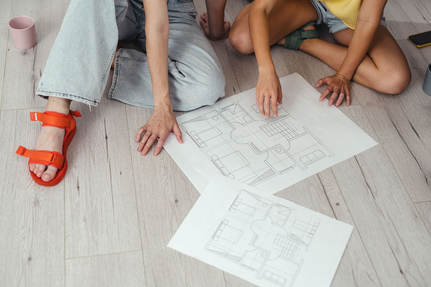 Two people sitting on a light wooden floor pointing at printed house floor plans