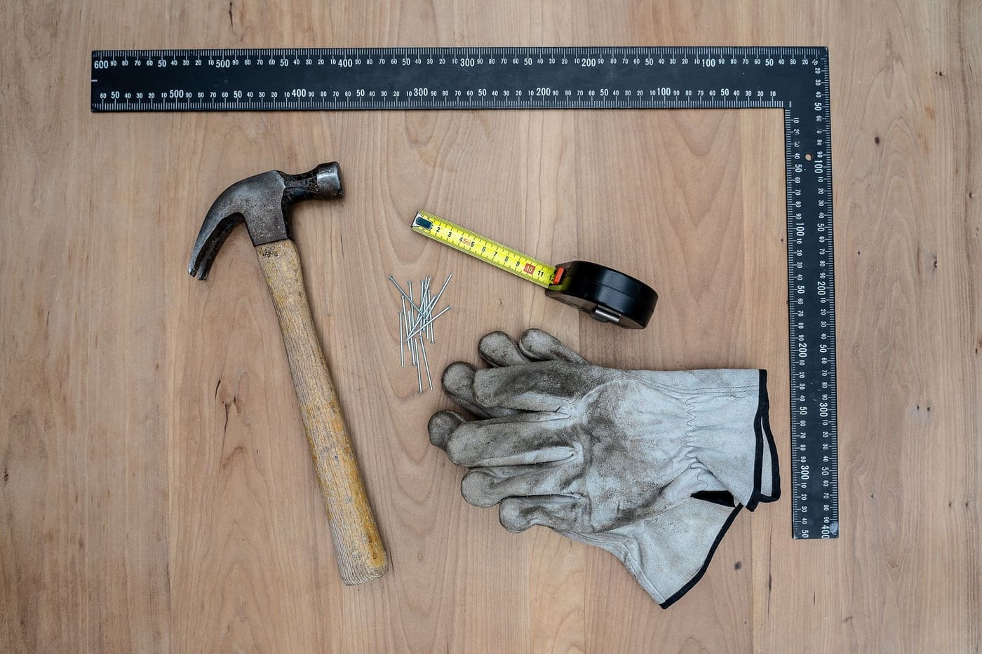 A hammer, tape measure, work gloves and a steel square laid out on a wooden surface