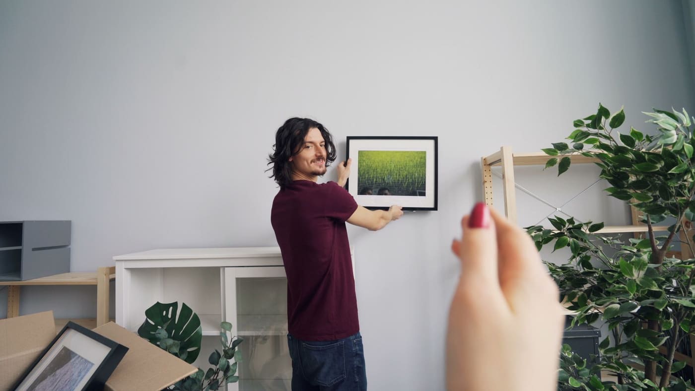 A man hanging a framed landscape picture on a living room wall beside a shelf and plants