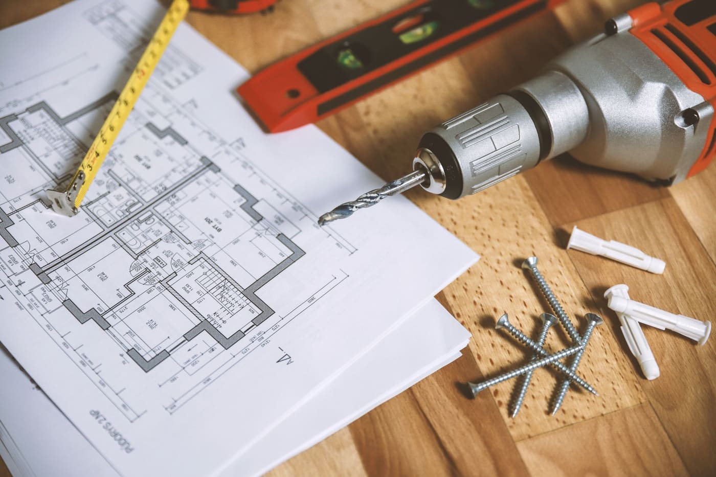 Printed floor plans on a wooden table with a tape measure, a drill, a level and wall anchors
