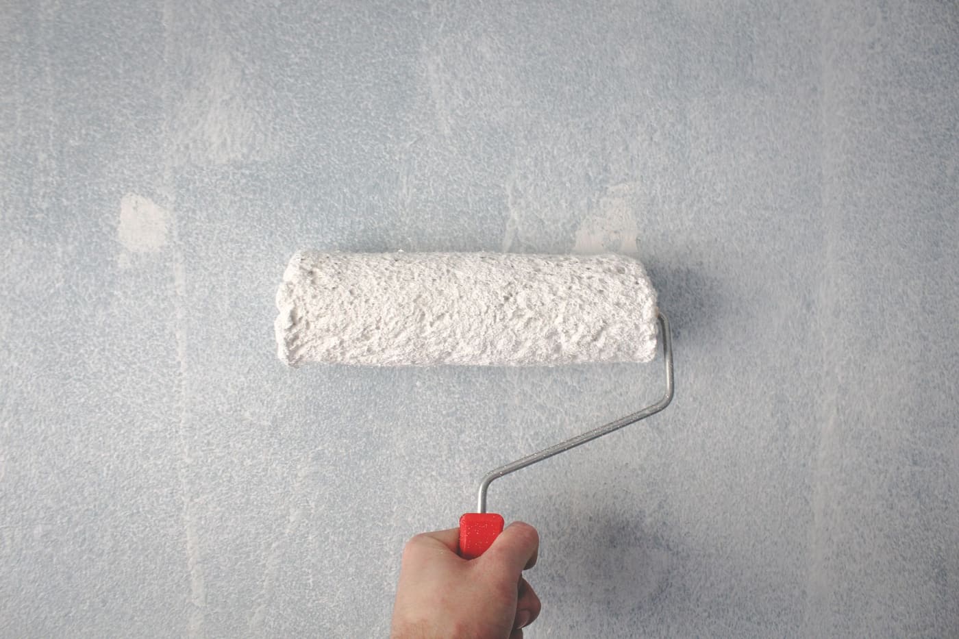 A hand holding a white paint roller against a grey wall with uneven coverage