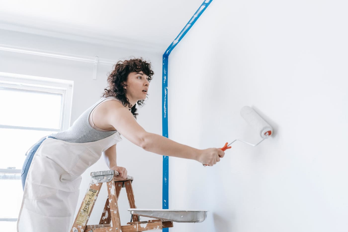 A woman rolling white paint onto a wall beside a blue taped edge, with a stepladder behind her