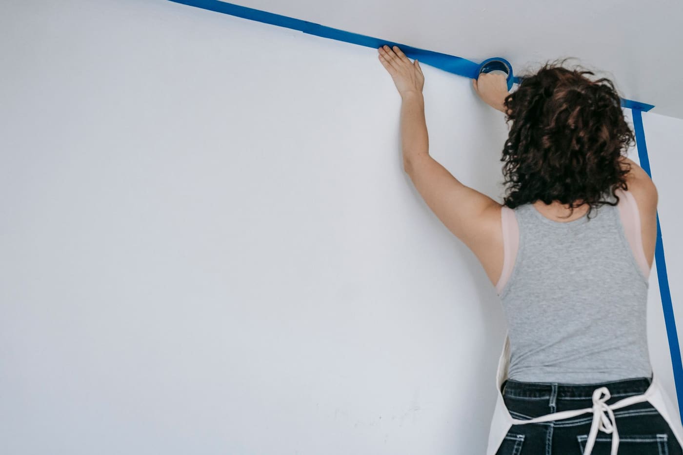 A woman pressing blue painter's tape along the ceiling line of a white wall